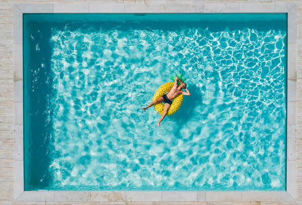 A cheerful middle-aged man in sunglasses suntanning floating on blue swimming pool waves on giant inflatable Yellow Pineapple tube and laughing. Chill out summer vacation in luxury resorts concept.