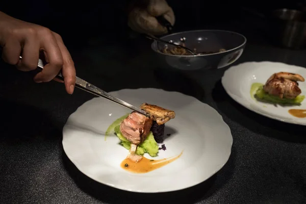 Chief cook putting Grilled meat piece using tweezers on green vegetables puree white plate with sauce. Healthy meals preparation, cooking or small business concept.