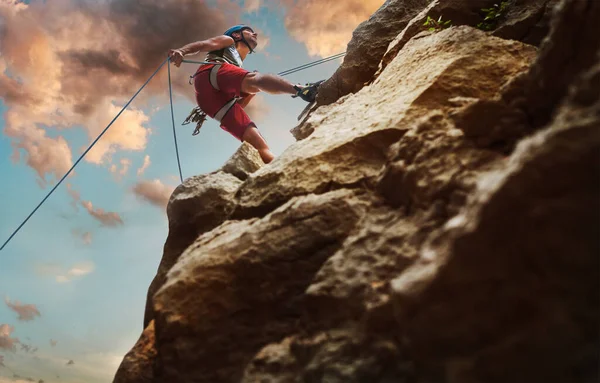 头戴防护头盔的肌肉攀登者用绳索系带装置从悬崖岩壁上垂下 并在黄昏的天空背景下攀爬 积极的极限运动时间概念 — 图库照片
