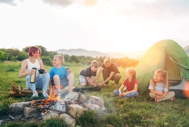 Bir grup gülümseyen çocuk dumanlı bir kamp ateşinin yanında neşeli bir konuşma yapıyor. Termostan çay içiyorlar, iki kardeş yeşil çadırı kuruyor. Mutlu aile piknik kampı konsepti.