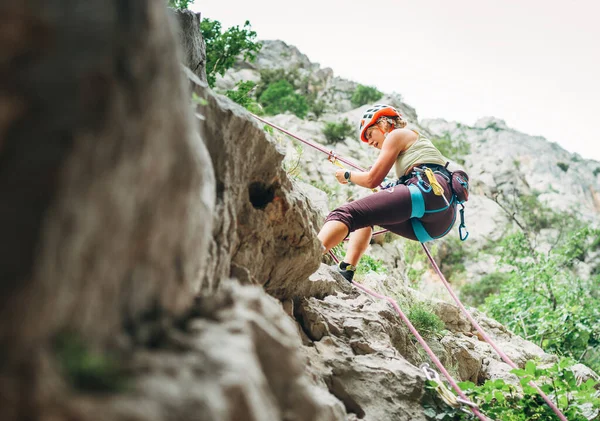 积极的登山者妇女戴着防护头盔 用绳索 腰带装置和攀爬安全带从悬崖岩壁上垂下 积极的极限运动时间概念 — 图库照片