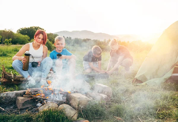一群笑着的孩子靠近一个冒烟的篝火边 喝着热水瓶里的茶 两个兄弟搭起了绿色的帐篷 快乐家庭户外野营活动的概念 — 图库照片