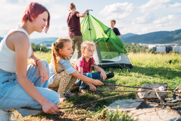 3人の姉妹はキャンプファイヤーの炎の上にマシュマロとキャンディーを笑って焙煎し 2人の兄弟は緑のテントを設置しました 幸せな家族の屋外ピクニックキャンプ活動のコンセプト — ストック写真