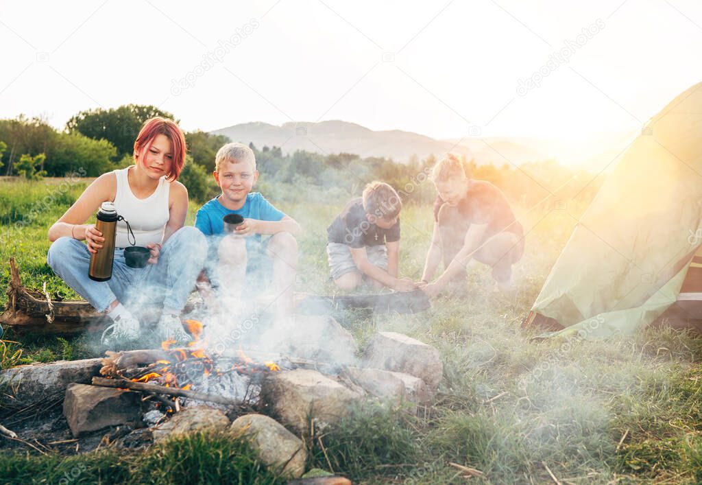 Group of smiling kids near a smoky campfire drinking tea from a thermos, two brothers set up the green tent. Happy family outdoor picnic camping activities concept