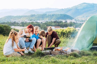 Bir grup gülümseyen çocuk dumanlı bir kamp ateşinin yanında neşeli bir konuşma yapıyor. Termostan çay içiyorlar, iki kardeş yeşil çadırı kuruyor. Mutlu aile piknik kampı konsepti.