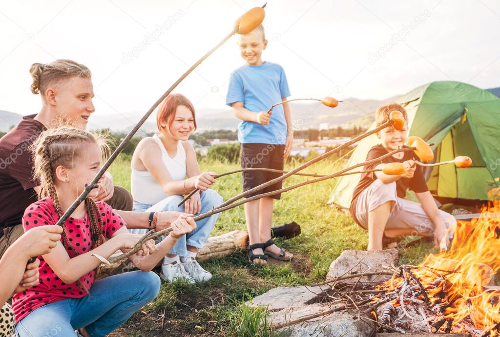 Group of six Kids: Boys and girls cheerfully smiling and roasting sausages on sticks over a campfire flame near the green tent. Outdoor active time spending or camping in Nature concept.