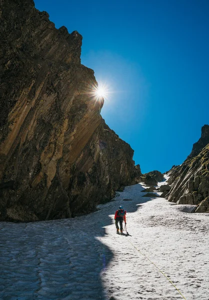 Team roping up woman with climbing axe dressed high altitude mountaineering clothes with backpack walking by snowy slope in the couloir with backlight sun on blue sky.