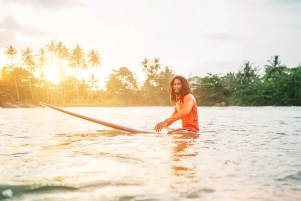 Black long-haired teen man floating on long surfboard, waiting for a wave ready for surfing with palm grove litted sunset rays. Extreme water sports and traveling to exotic countries concept.