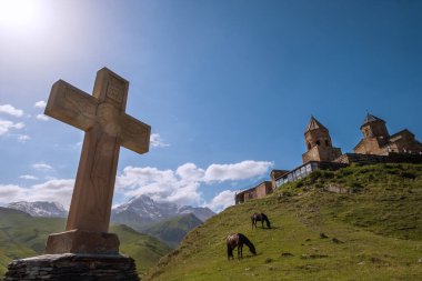 Yeşil çim tepesi ve Kazbek 5054 metre dağında otlayan atlarla Gergeti Trinity Kilisesi ile taş Ortodoks haçı. Gürcistan 'ın Stepantsminda köyü yakınlarındaki geniş açı manzarası.