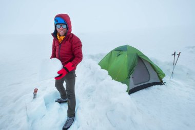 Gülümseyen adam, kırmızı, sıcak dağları aşan bir ceket giyerek Kazbek Dağı 'nın 4200 metre altındaki buzullu platodaki kamp çadırının etrafındaki kar tuğlalarından koruyucu duvarı örüyor. Doğu Kafkas Dağları, Gürcistan.