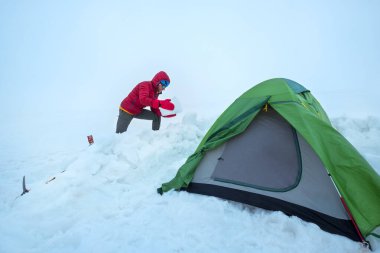 Gülümseyen adam, kırmızı, sıcak dağları aşan bir ceket giyerek Kazbek Dağı 'nın 4200 metre altındaki buzullu platodaki kamp çadırının etrafındaki kar tuğlalarından koruyucu duvarı örüyor. Doğu Kafkas Dağları, Gürcistan.