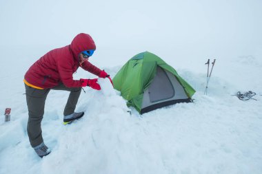 Gülümseyen adam, kırmızı, sıcak dağları aşan bir ceket giyerek Kazbek Dağı 'nın 4200 metre altındaki buzullu platodaki kamp çadırının etrafındaki kar tuğlalarından koruyucu duvarı örüyor. Doğu Kafkas Dağları, Gürcistan.