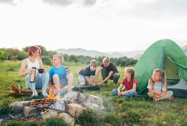笑顔の子供たちのグループは 煙の多いキャンプファイヤーの近くで陽気な会話をしています 彼らは魔法瓶からお茶を飲む 2人の兄弟は緑のテントを設定します 幸せな家族の屋外ピクニックキャンプ活動のコンセプト — ストック写真