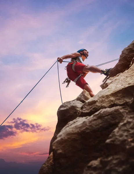 头戴防护头盔的肌肉攀登者用绳索系带装置从悬崖岩壁上垂下 并在黄昏的天空背景下攀爬 积极的极限运动时间概念 — 图库照片