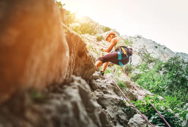 积极的登山者妇女戴着防护头盔 用绳索 腰带装置和攀爬安全带从悬崖岩壁上垂下 积极的极限运动时间概念 — 图库照片