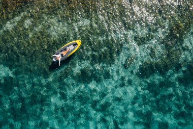 A lonely female in a straw hat smiling, relaxing lying floating in a kayak on the turquoise Adriatic Sea waves. Aerial coastal top view shot. Exotic countries vacations concept.