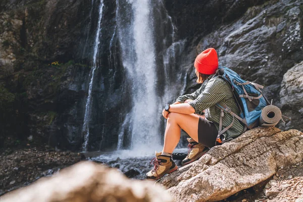 坐在山河瀑布边 头戴红色礼帽 身穿活跃的徒步服的女人 享受着大自然的神奇力量 远足和自然概念形象 — 图库照片