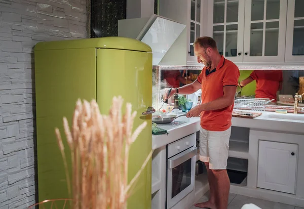 明るい緑の冷蔵庫で居心地の良い家庭のキッチンで朝の朝食を調理笑顔 彼は鍋でおいしいものを揚げている 健康的な食事の準備と人々のライフスタイルのコンセプトイメージ — ストック写真