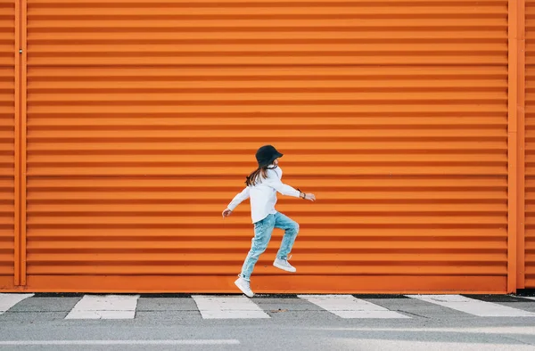 穿着时髦的小女孩在橙色墙壁的背景上跳过一条人行横道 这很滑稽 城市居民的生活与街头的日常生活观念形象 — 图库照片