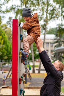 Küçük çocuk babasının desteğiyle çocuk parkında kaya duvarına tırmanıyor.