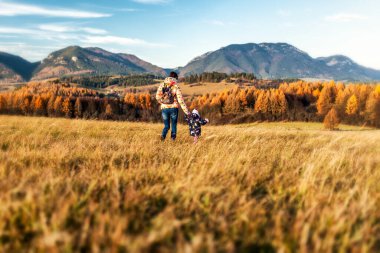 Anne çocuğuyla birlikte çayırda yürüyor ve bebeğinin elini tutuyor. Arka planda sonbahar ülkesi.