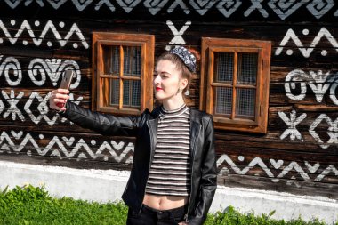 Siyahlı genç kız Slovakya 'nın kırsal kesimindeki Cicmany köyünün eski kır evinin önünde selfie çekiyor.