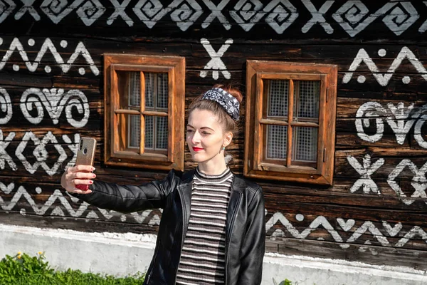 Siyahlı genç kız Slovakya 'nın kırsal kesimindeki Cicmany köyünün eski kır evinin önünde selfie çekiyor.