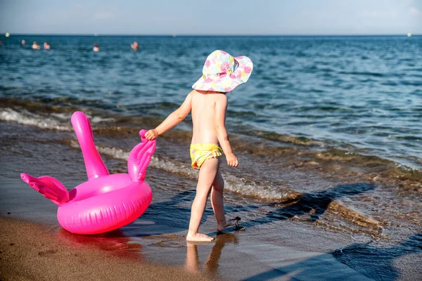 Small child with inflatable pink flamingo walking into the sea.