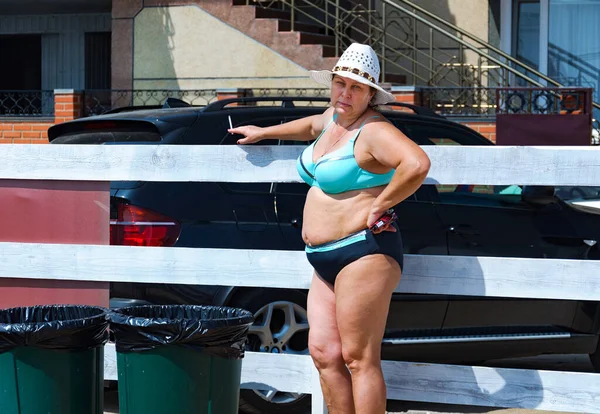 Bikini giymiş tombul bir kadın sahildeki sigara içme alanında çitin yanında sigara içiyor..