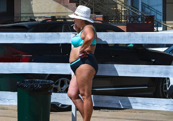 Bikini giymiş tombul bir kadın sahildeki sigara içme alanında çitin yanında sigara içiyor..