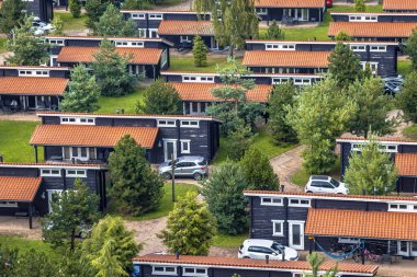 Yeşil ormanlık alanda benzer simetrik sırayla yazlık evleri olan Holiday Bungalow Parkı. Çimlerde ve çalılarda turuncu çatı kiremitleri olan ahşap kiremitler. Hollanda.