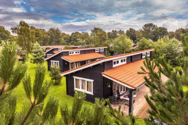 Yeşil ormanlık alanda benzer simetrik sırayla yazlık evleri olan Holiday Bungalow Parkı. Çimlerde ve çalılarda turuncu çatı kiremitleri olan ahşap kiremitler. Hollanda.