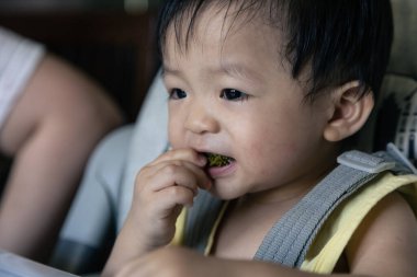 Sevimli, neşeli ve mutlu 1 yaşında Çinli bir çocuk bebek koltuğunda otururken yemek yiyor.