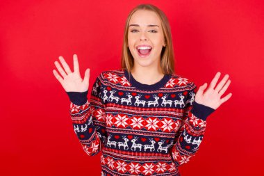 İyimser genç Kafkasyalı kız Noel süveteri giyiyor, sevinçten avuçlarını kaldırıyor, birinden müthiş bir hediye almaktan mutlu, yüksek sesle bağırıyor, heyecanlı manken çığlığı atıyor..