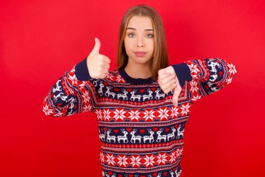 Kafkasyalı genç kız Noel süveteri giyip baş parmağını kaldırıyor, zor seçim konsepti.