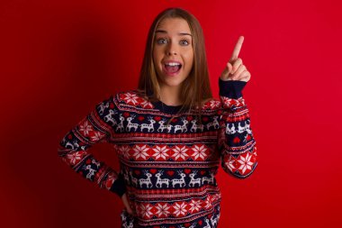 Kafkasyalı genç kız Noel süveteri giyiyor, parmağını kaldırıyor, fikir yürütüyor ve poz veriyor.
