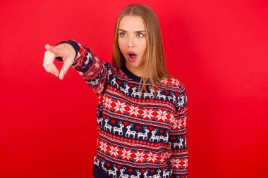 Kafkasyalı genç kız noel süveteri giyiyor. Parmağıyla işaret ediyor. İleriye şaşırmış, ağzı açık..