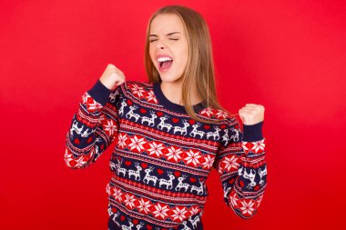 Kırmızı arka planda Noel süveteri giyen genç beyaz kız kolları havada ve gözleri kapalı bir şekilde başarıyı kutluyor ve şaşırıyor. Kazanan konsept.