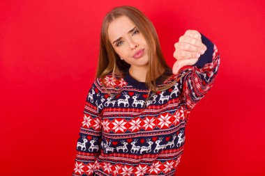 Kırmızı arka planda Noel süveteri giyen genç beyaz bir kız kızgın, sinirli, hayal kırıklığına uğramış ya da hoşnutsuz, ciddi bir bakışla başparmaklarını göstermiyor.