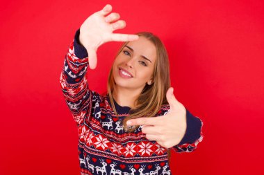 Kırmızı arka planda Noel süveteri giyen beyaz bir kız elleriyle parmak çerçevesi yapıyor. Yaratıcılık ve fotoğrafçılık kavramı.