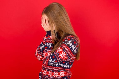 Üzgün Kafkasyalı bir kız kırmızı arka planda Noel süveteri giyiyor ve elleriyle yüzünü kapatarak ağlıyor..