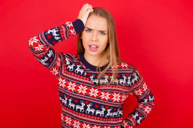 Utanmış, Kafkasyalı genç bir kız kırmızı arka planda Noel süveteri giyiyor, şok olmuş bir ifadeyle, büyük şaşkınlık içinde, bilmece gibi, kapalı alanda poz veriyor.