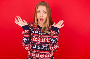 Şaşırmış genç beyaz kız kırmızı arka planda Noel süveteri giyiyor belirsizlikle jestler yapıyor, kameraya bakıyor, şaşırmış bir şekilde hileli soruya cevap vermiyor, insanlar, vücut dili, duygu kavramı.