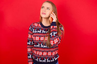 Üzgün genç beyaz kız kırmızı arka planda Noel süveteri giyip akıllı telefondan konuşuyor. İletişim konsepti.