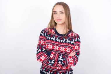 Kendine güveni tam, beyaz arka planda Noel süveteri giyen, kolları bağlı, sakin, beyaz bir kız. Profesyonel titreşimlerin kendine güvenen pozlarda durduğunu gösterir.