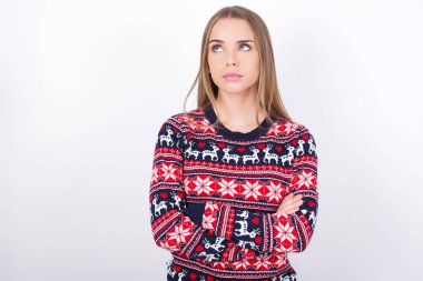 Beyaz arka planda Noel süveteri giyen düşünceli genç beyaz bir kız kollarını kavuşturmuş düşünceli bir ifadeyle ne yapacağını düşünüyor.
