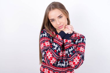 Beyaz arka planda Noel süveteri giyen beyaz tenli çekici genç beyaz kız kameraya gülümseyerek gülümsüyor.