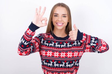 Beyaz arka planda Noel süveteri giyen genç beyaz bir kız gülümsüyor ve mutlu görünüyor, kaygısız ve pozitif, bir eliyle zafer ya da barış işareti yapıyor.