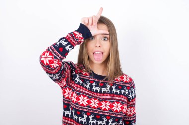 Beyaz arka planda Noel süveteri giyen, alnında parmak olan genç beyaz bir kız ezikçe bir jest yapıyor. İnsanlar dil gösteriyor.