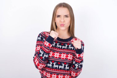 Displeased annoyed Young caucasian girl wearing christmas sweaters on white background clenches fists, gestures pissed, ready to revenge, looks with aggression at camera stands full of hate, being pressured